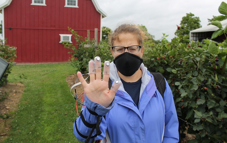 A woman in a face mask raises her hand, signaling or greeting, with a focused expression on her face.