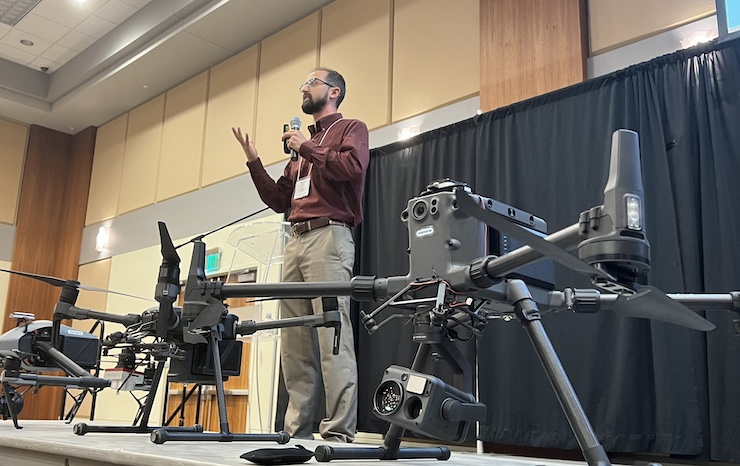 A man stands confidently in front of a large drone, showcasing its impressive size and advanced technology.
